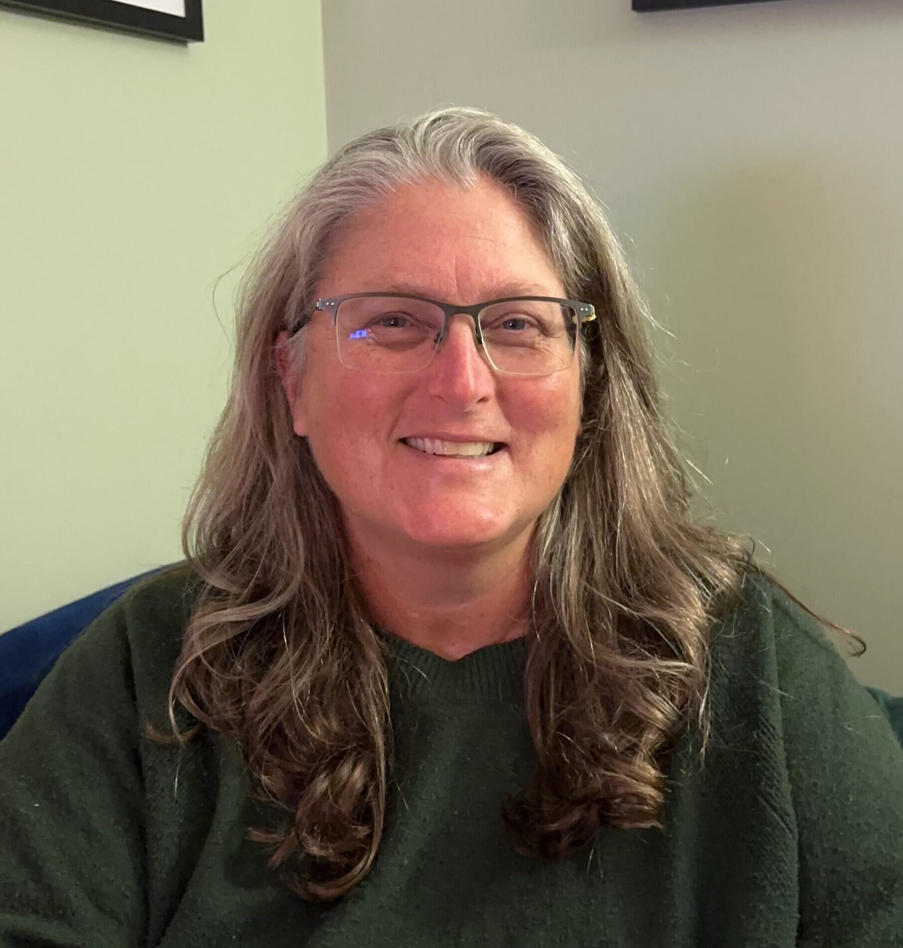 Regan Brashear, somatic coach with Welcome Home Somatics, sitting on green couch, smiling at the camera.