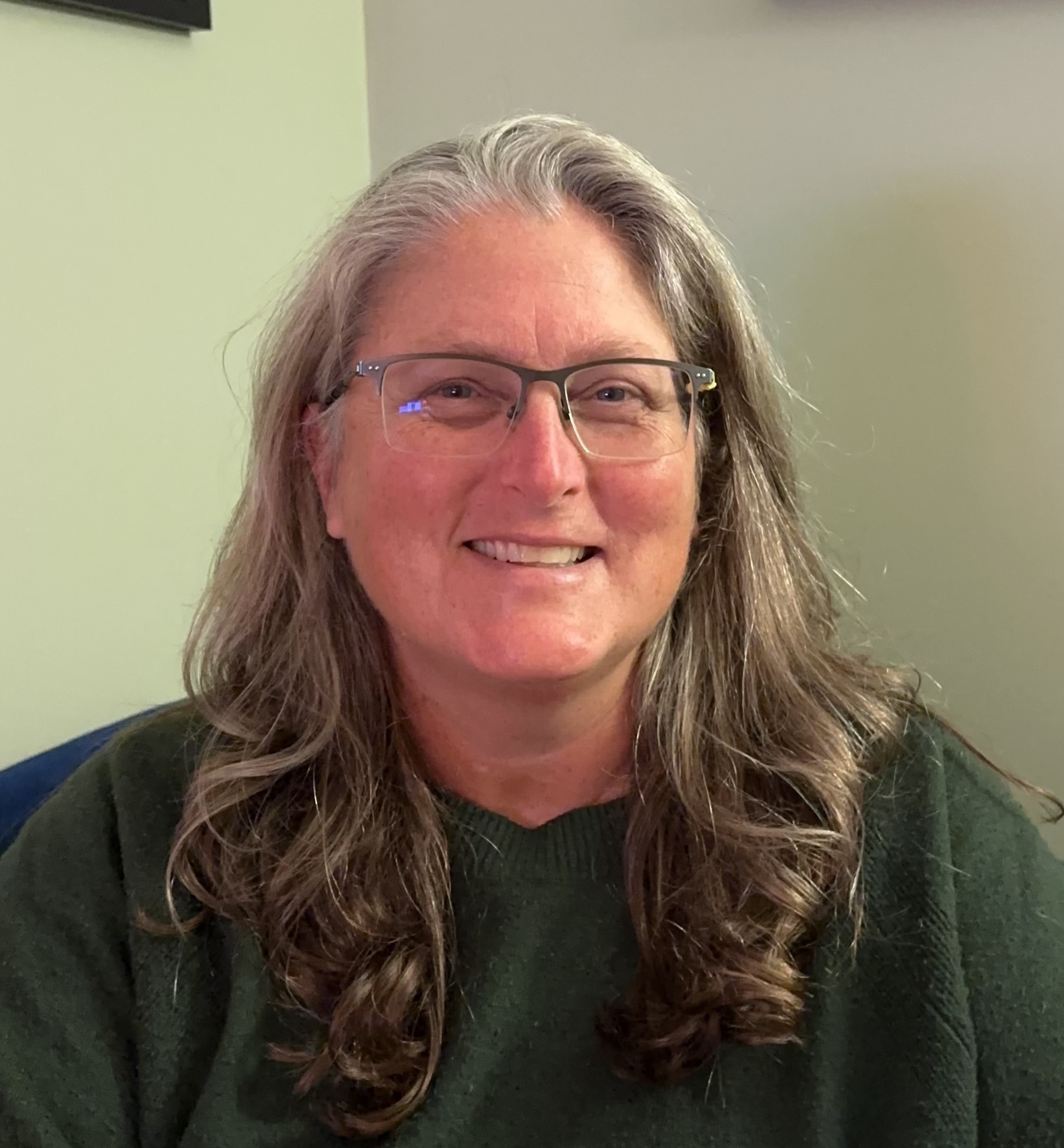 Regan Brashear, somatic coach with Welcome Home Somatics, sitting on green couch, smiling at the camera.