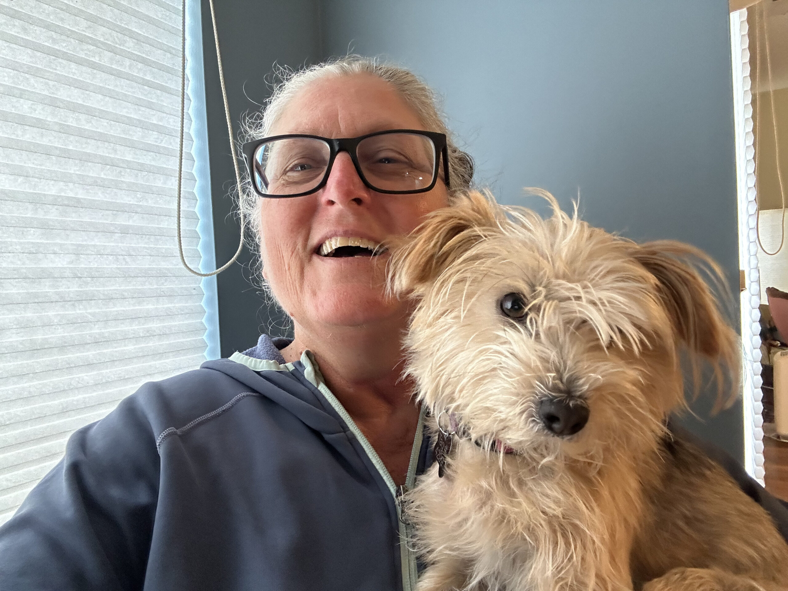 Image of white person with black glasses and a big open-mouthed grin holding a scruffy sandy-colored dog in their lap